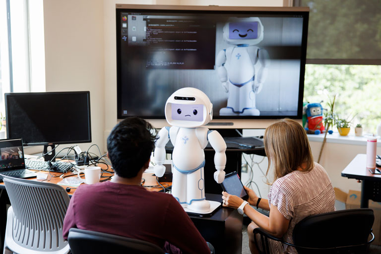 Man and a woman with a robot sitting on a table in front of them, as they write code to make the robot work.