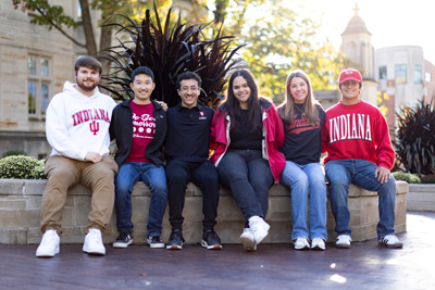 6 Luddy Student Ambassadors sitting on a rock wall with the IU Campus in the background.