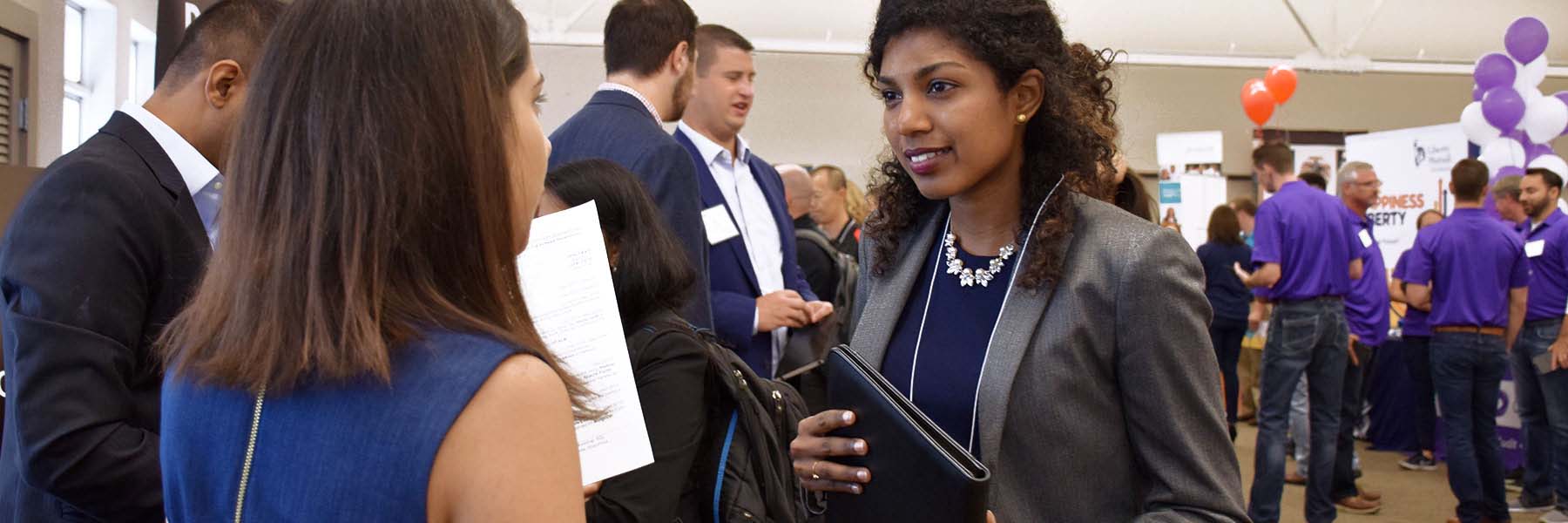 A student holds a portfolio and speaks with a representative at a career fair.