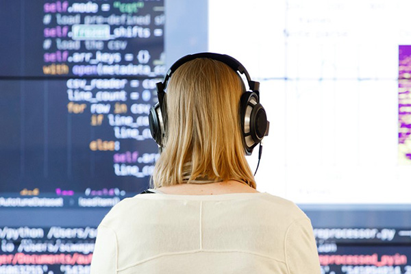 a girl with headphones sitting in front a big screen with code on it.