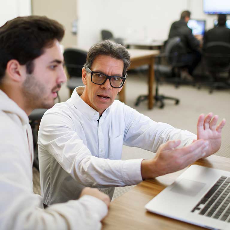 A male speaks to a male colleague while looking at a laptop. 