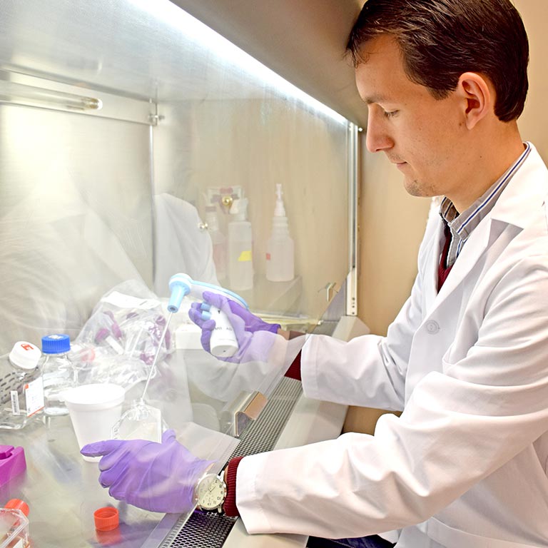 A researcher in a lab coat uses equipment to fill a container with liquid behind a screen.